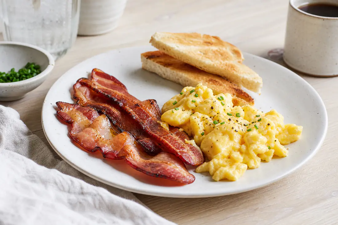 Sprøstekte baconskiver på en hvit tallerken med eggerøre og ristet brød