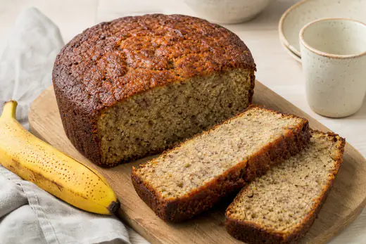 Rundt bananbrød med mørk skorpe og to tykke skiver skåret ut, på et trebrett med en moden banan ved siden av