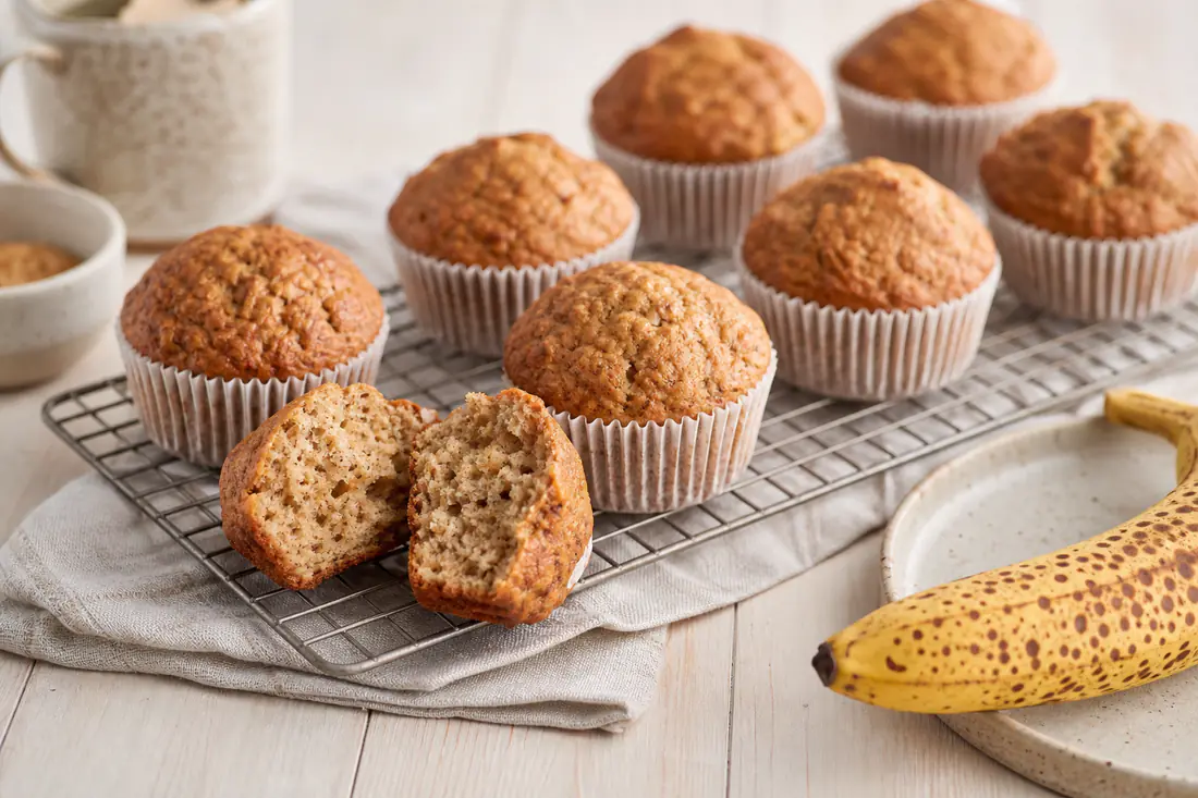 Gylne bananmuffins på en rist med en moden banan og en muffins delt i to ved siden av