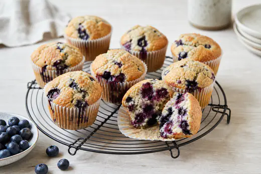 Blåbærmuffins i papirformer på en rist, med en oppskåret muffins som viser saftige blåbær inni