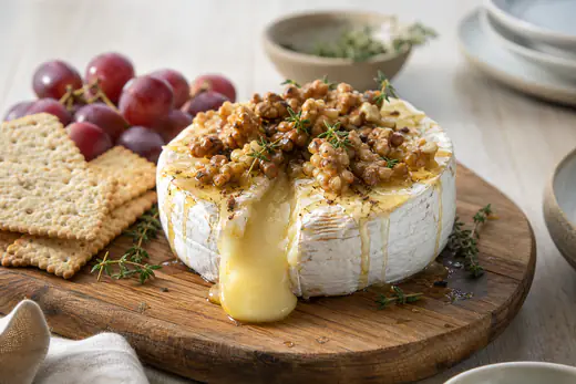 Hel bakt brie med smeltet midte, honning, valnøtter og timian på et trebrett med kjeks og druer