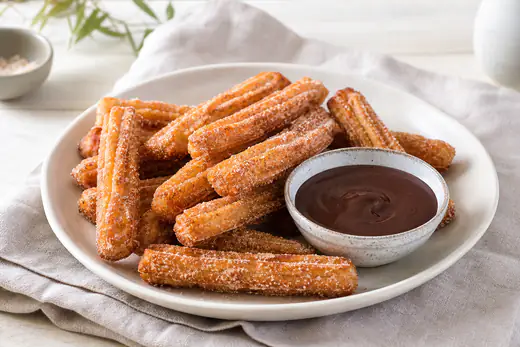 Riflede gyllenbrune churros rullet i kanelsukker på en tallerken med en skål varm sjokoladesaus