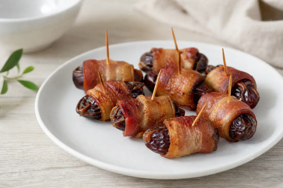 Dadler pakket inn i sprø, gyllen bacon på et lite fat med tannstikkere og et glass rødvin ved siden av