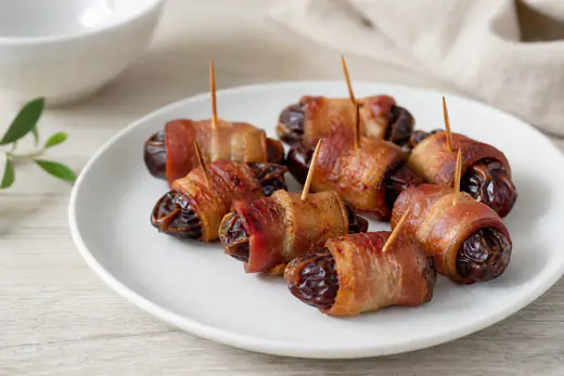 Dadler pakket inn i sprø, gyllen bacon på et lite fat med tannstikkere og et glass rødvin ved siden av