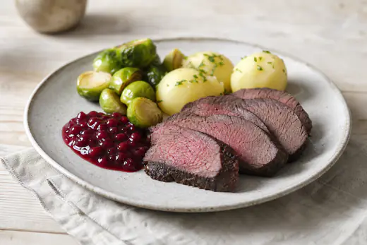 Skåret elgstek med rosa kjøtt og mørk skorpe på et fat med rosenkål, poteter og tyttebærsyltetøy