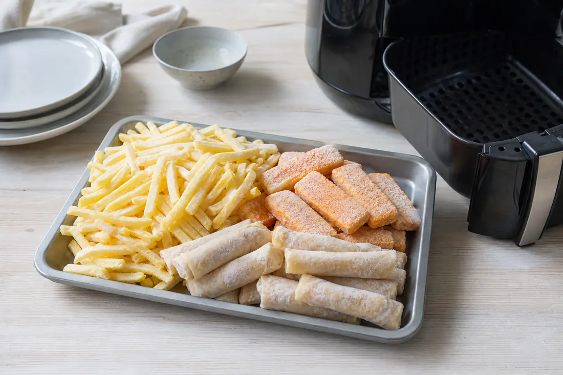 Frosne pommes frites, fiskepinner og vårruller med rim på et brett ved siden av en airfryerkurv