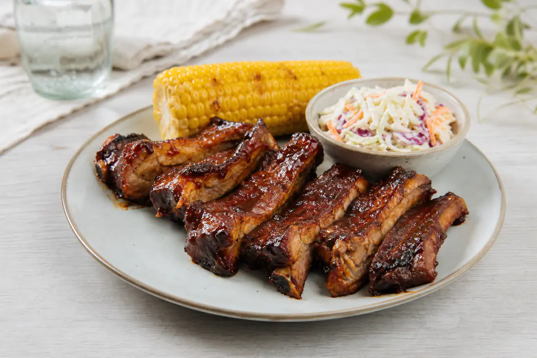 Blanke, mørkebrune spareribs med glaze på en tallerken med maiskolbe og coleslaw