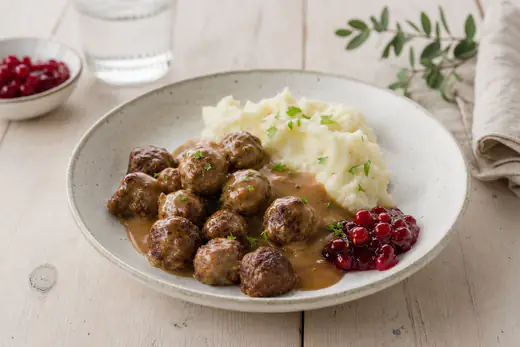 Små brunstekte kjøttboller i en skål med brun saus, potetmos og tyttebærsyltetøy ved siden av
