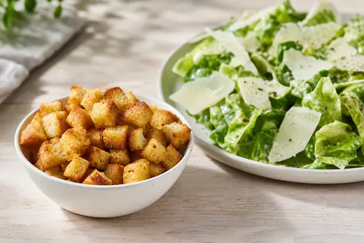 Skål med gyldne, sprø brødterninger som krutonger, og en grønn salat med parmesan ved siden av