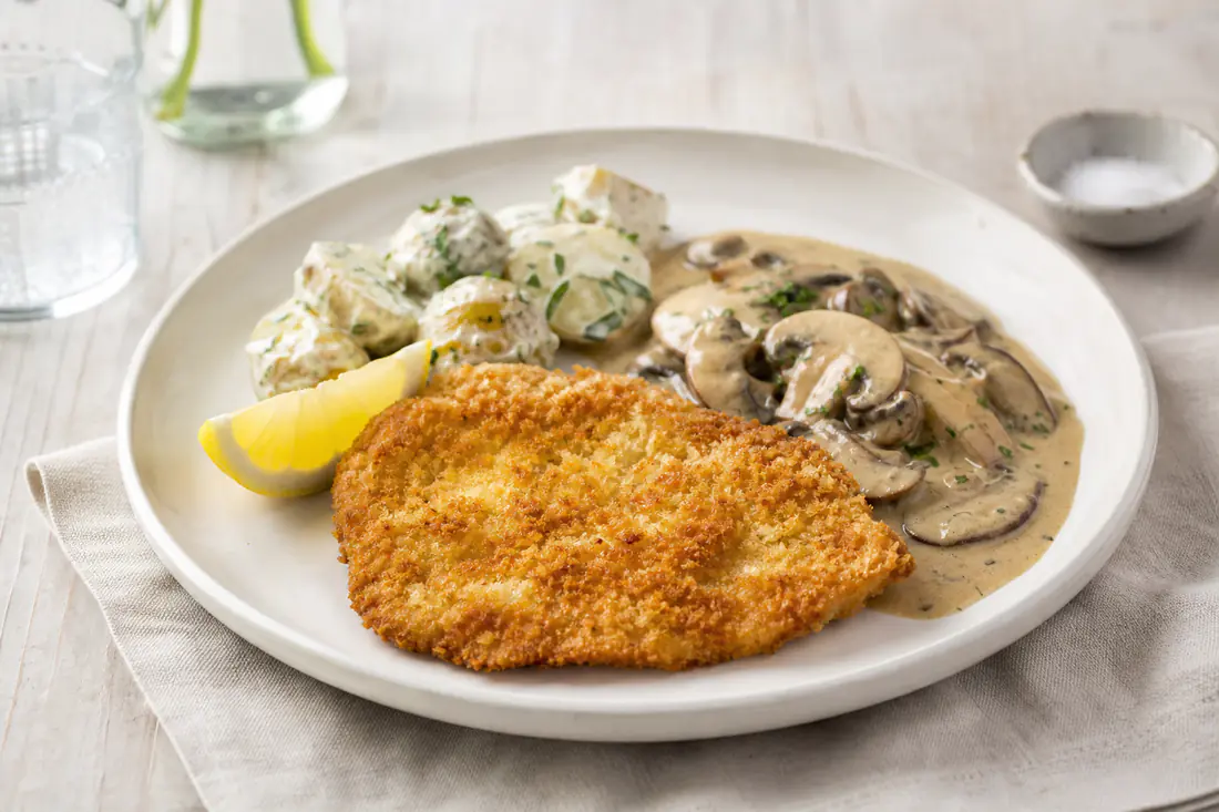 Gyllen, panert kyllingschnitzel på en tallerken med sitronbåt, kremet soppsaus og potetsalat