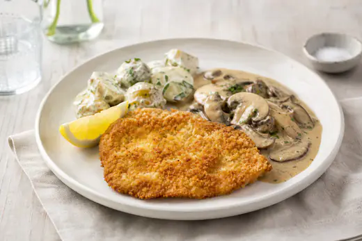 Gyllen, panert kyllingschnitzel på en tallerken med sitronbåt, kremet soppsaus og potetsalat