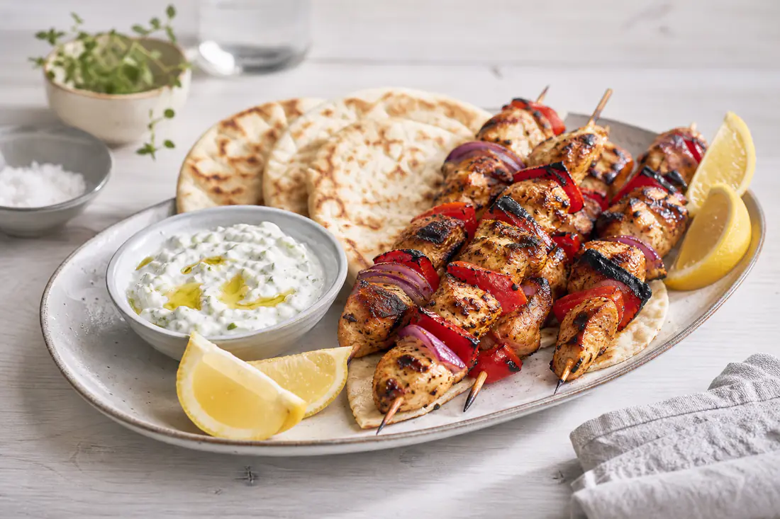 Grillede kyllingspyd med biter av rød paprika og rødløk på et fat med tzatziki, pitabrød og sitronbåter