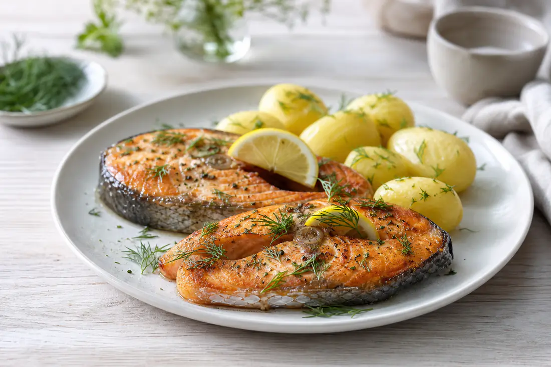 To gylne laksekoteletter med ben, toppet med frisk dill og sitronbåter, på en tallerken med kokte poteter