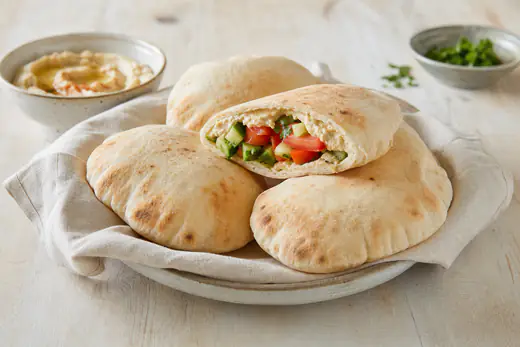 Stabel med hjemmebakte, oppblåste pitabrød og ett pitabrød delt i to med hummus og agurk