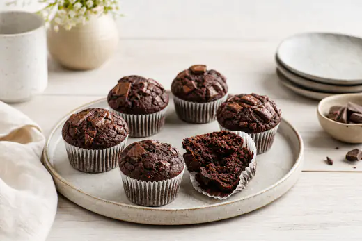 Seks mørke sjokolademuffins med sjokoladebiter på toppen i papirformer på et brett, en av dem delt i to