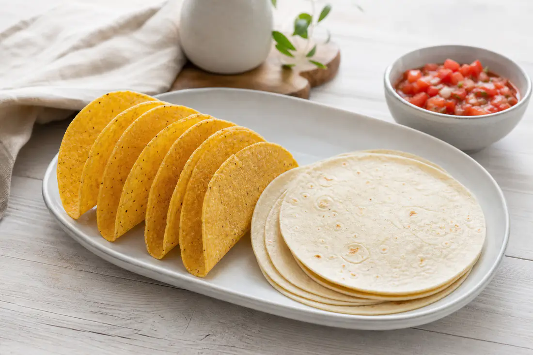 Sprø, gyllenbrune tacoskjell av tortilla stående på et brett ved siden av varme, brettede tortillalefser